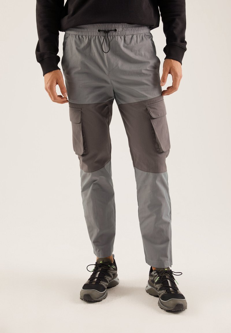 Pantalons cargo gris et gris foncé avec une taille élastique, cordon de serrage ajustable et poches latérales. Fabriqués en tissu léger et lisse.