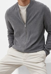 Cardigan grigio con zip e colletto tondo; texture a maglia morbida; dettaglio della zip argentata. Indossato sopra una t-shirt chiara, abbinato a pantaloni beige.