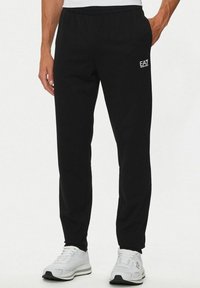 Uomo che indossa pantaloni neri EA7 Emporio Armani e sneakers bianche, in piedi con una mano in tasca su uno sfondo neutro.