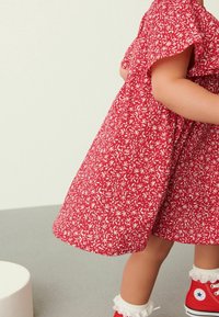 Ein Kind trägt ein rotes Blumenkleid, weiße Rüschensocken und rote High-Top-Sneaker und steht auf einem grauen Boden neben einem weißen zylindrischen Objekt.