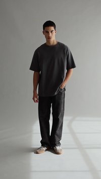 Ung man står med en hand i fickan, iförd en löst sittande mörkgrå t-shirt, svarta byxor och tan sneakers i en minimalistisk inomhusmiljö.