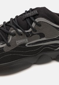 Black sneaker featuring a textured mesh upper, grey accents, and a prominent CAMPER logo. The sole is thick with a wave-like pattern.