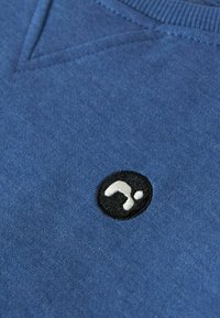 Blauwe trui met v-hals, zachte textuur en een klein zwart rond geborduurd logo aan de onderzijde.