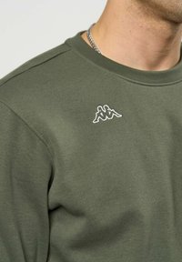 Nærbilde av en person som har på seg en olivengrønn genser med en liten hvit logo av to sittende figurer rygg mot rygg på brystet.