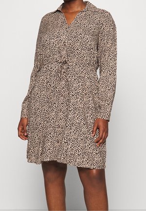 Femme portant une robe à manches longues à imprimé léopard beige et noir, avec un col et une taille à cordon, debout devant un fond uni.