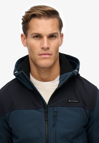 Marineblaue Zip-Jacke mit einer kontrastierenden dunkelblauen Kapuze und einem Kragen, ausgestattet mit einer strukturierten Oberfläche und einem sichtbaren Logo auf der Brust.