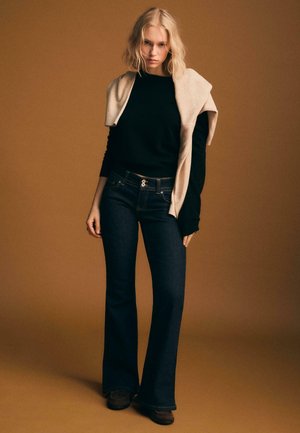 Czarny sweter z długim rękawem w warstwach z beżowym płaszczem, zestawiony z ciemnoniebieskimi rozszerzanymi dżinsami. Model nosi brązowe buty, na tle brązowego tła.