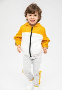 Ungdoms löpningsoutfit med en gul och vit zip-up jacka med elastiska manchetter och grå joggers med gula sidränder. Vita sneakers.