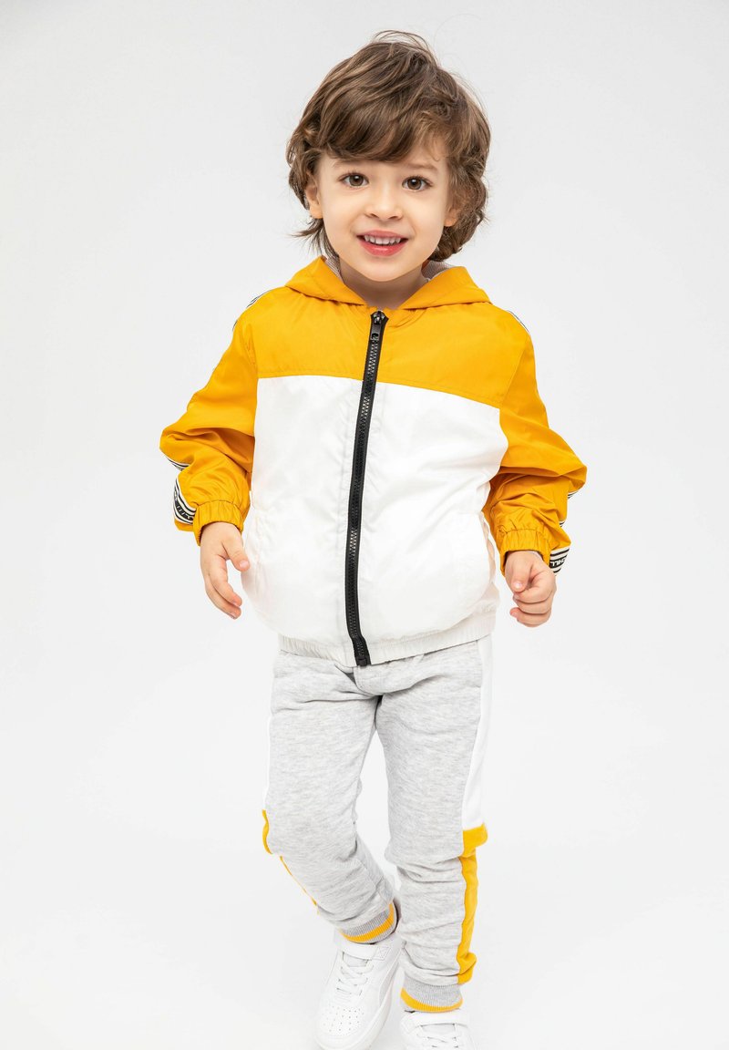 Ungdoms löpningsoutfit med en gul och vit zip-up jacka med elastiska manchetter och grå joggers med gula sidränder. Vita sneakers.