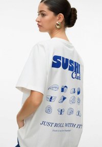 Biała, oversize koszulka z niebieskimi grafikami sushi oraz napisem „SUSHI Club” i „JUST ROLL WITH IT!” na plecach. Krótkie rękawy, casualowy krój.