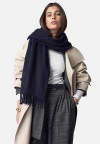 Mørkeblå uldhalstørklæde med frynser draperet over en lys beige trenchcoat, båret over en hvid top og grå mønstrede bukser.