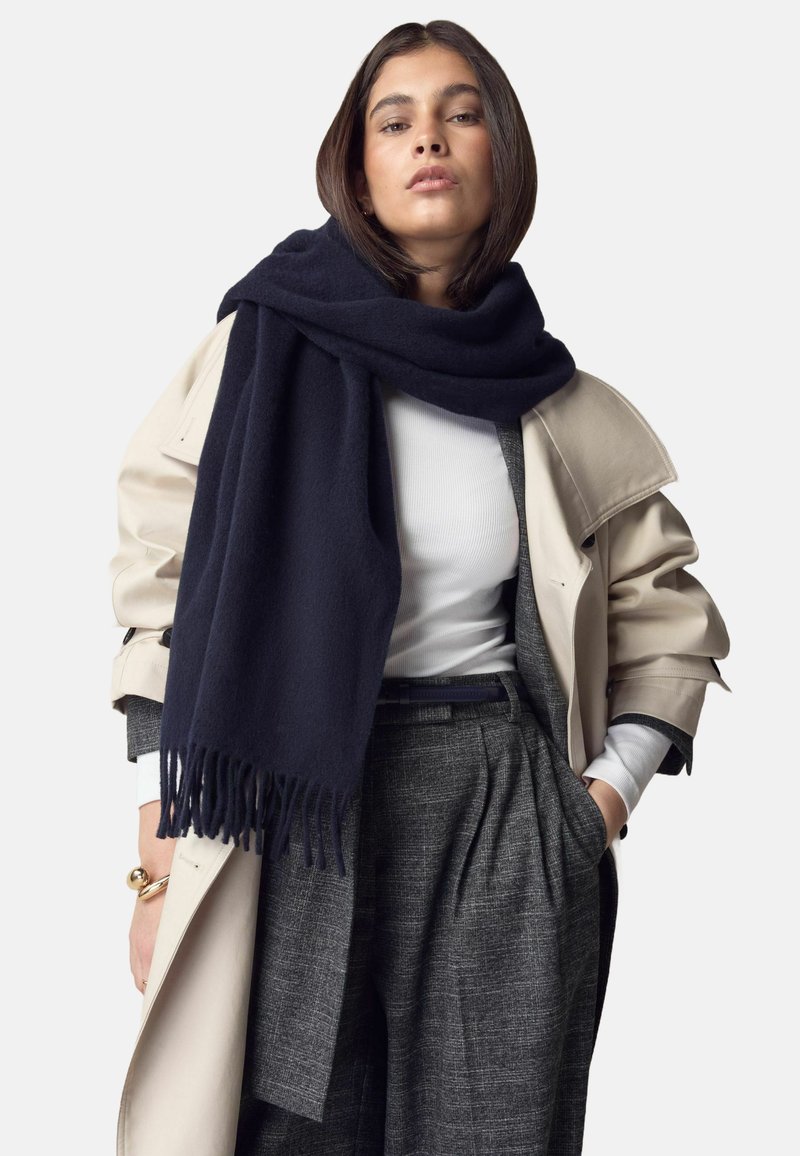 Mørkeblå uldhalstørklæde med frynser draperet over en lys beige trenchcoat, båret over en hvid top og grå mønstrede bukser.