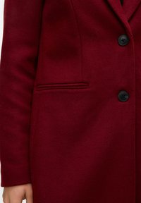 Cappotto di lana bordeaux con colletto a bavero, design elegante, tre bottoni neri e una tasca laterale con dettaglio a piping stretto.