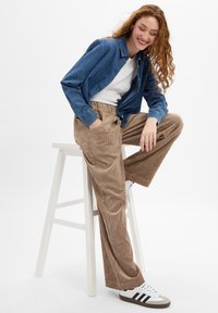 Chemise en denim bleue, T-shirt blanc en dessous, associée à un pantalon large en velours côtelé marron et des baskets blanches avec des rayures noires. Assise sur un tabouret.