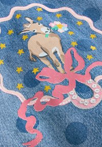 Tissu en denim avec un design fantaisiste mettant en scène une chèvre brune, des fleurs colorées, des rubans roses et des étoiles jaunes sur fond bleu.