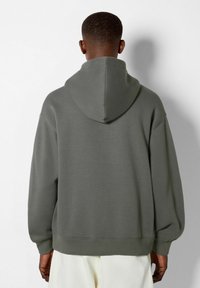 Uomo che indossa una felpa con cappuccio verde oliva, di spalle, mostrando i dettagli posteriori e il taglio morbido sopra dei pantaloni bianchi.