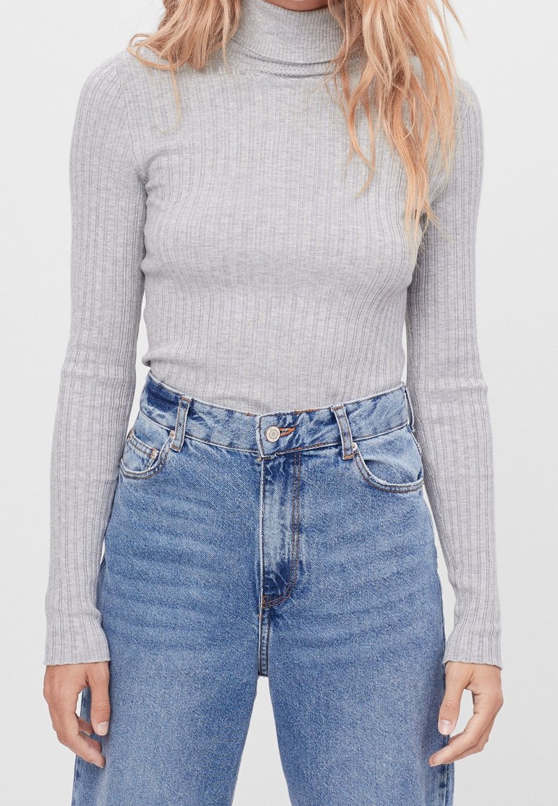 Personne portant un pull col roule côtelé gris clair rentré dans un jean en denim bleu taille haute, se tenant devant un fond uni.