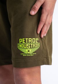 Shorts vert olive en tissu doux avec un grand logo graphique jaune vif portant l'inscription "PETROL INDUSTRIES" avec des accents ailés.