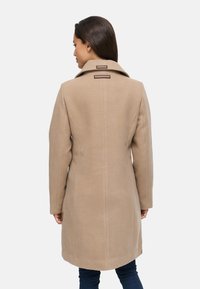 Cappotto di lana beige con un design strutturato, caratterizzato da un alto colletto, cuciture sottili e due etichette di marca vicino al colletto sul retro.