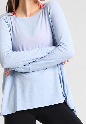 Kobieta w jasnoniebieskiej koszulce z długim rękawem na różowym biustonoszu sportowym, z założonymi skrzyżowanymi ramionami, ubrana w czarne legginsy na tle jednolitego tła.