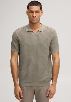 Homme portant un polo à manches courtes texturé gris clair et un pantalon assorti, debout devant un fond clair uni.