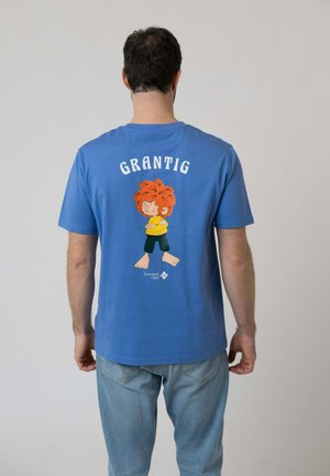 Mann trägt blaues T-Shirt mit einem Cartoon-Figur mit roten Haaren und dem Wort "GRANTIG" auf der Rückseite, kombiniert mit hellblauen Jeans.