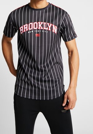 Mann trägt ein schwarzes Nadelstreifen-T-Shirt mit der Aufschrift "BROOKLYN New York City" in roten und weißen Buchstaben, kombiniert mit schwarzen Hosen.