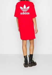 Czerwona, oversize'owa sukienka t-shirt z białym logo adidas i wzorem trefoil na plecach. Materiał wydaje się gładki. Połącz z czarnymi butami.