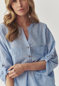 Camicia azzurro chiaro, con bottoni e colletto, due tasche sul petto e maniche arrotolate. Realizzata in un tessuto morbido e leggero.