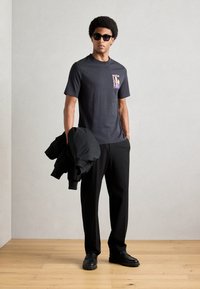 Sort bomulds T-shirt med et grafisk motiv på venstre bryst, parret med sorte bukser og en sort jakke, som holdes i den ene hånd. Solbriller båret.
