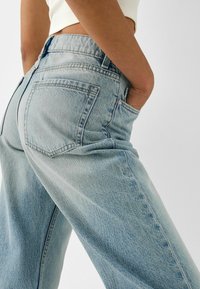 Világoskék, magas derekú farmernadrág, kényelmes fazonnal, hátsó zsebekkel és kopott denim textúrával. Oldalnézet, amely a csípő részleteit mutatja.