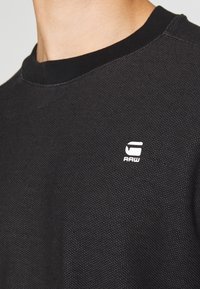 Černé texturované tričko s crewneck límcem a malým bílým logem "G RAW" na horní části hrudníku blízko ramene.