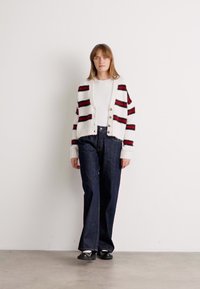 Cardigan a righe in bianco, rosso e nero, con chiusura a bottoni e polsini a coste, indossato sopra una maglietta bianca e jeans in denim scuro a vita alta.