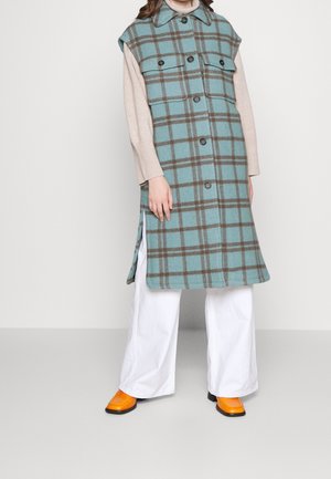 Personne portant un long gilet à carreaux bleu sarcelle et marron sur un pull beige, un pantalon large blanc et des chaussures orange vif à bout carré.