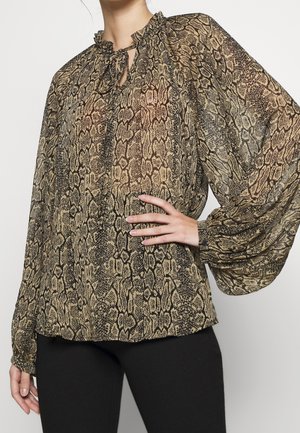 Vrouw draagt een blouse met slangenprint, lange mouwen, ruches aan de kraag en een striksluiting, gecombineerd met een zwarte broek, hand op de heup.