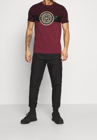 Bordeaux t-shirt met een zwarte patroon bovenkant en een ronde gouden logo. Gecombineerd met zwarte cargobroek en zwarte sneakers. Matte en gestructureerde stoffen.