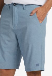 Blue short pants made of textured fabric, featuring a button closure, side pockets, and a small logo patch near the hem.