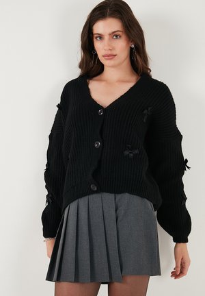 Cardigan noir en tricot avec un col en V, comportant cinq boutons noirs et des nœuds décoratifs sur les manches. Associé à une jupe grise plissée.