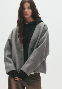 Siva jakna iz volnenega mešanega materiala s sproščenim fitom, ki ima dva gumba, široke rokave in temne manšete. Nosi se čez črn kapuco.