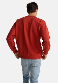 Felpa rossa con collo rotondo, maniche lunghe e polsini a coste, abbinata a jeans di un blu chiaro. Materiale in cotone con una consistenza liscia.