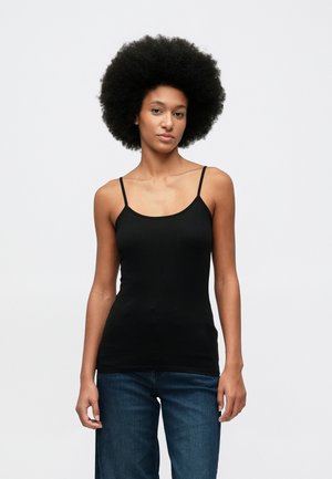 Jeune femme avec une coiffure afro naturelle, portant un top noir sans manches et un jean bleu, se tenant devant un fond blanc uni.