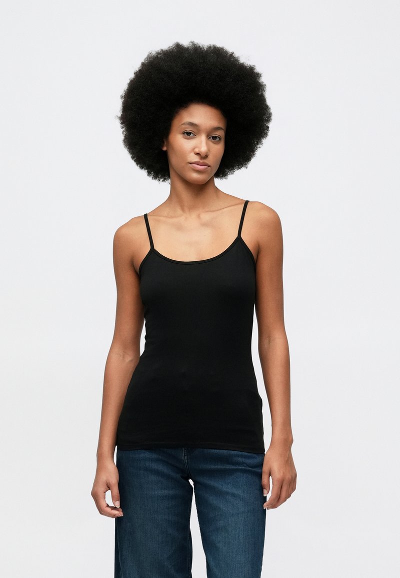 Jonge vrouw met een natuurlijke afro-haarstijl, gekleed in een zwarte mouwloze top en blauwe jeans, staande tegen een eenvoudige witte achtergrond.