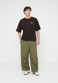 Μαύρο βαμβακερό μπλουζάκι με πράσινο λογότυπο, συνδυασμένο με φαρδιά ελαιί πράσινη παντελόνια με κίτρινα ραφές και λεπτομέρειες, φορεμένο με λευκά αθλητικά παπούτσια.