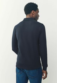 Camisa polo de manga larga en navy con superficie texturizada, cuello acanalado y aberturas laterales, combinada con jeans de mezclilla azul. Se muestra vista trasera.