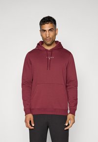 Maroonsweater med en frontlomme, snoretræk i huen og hvidt logo på brystet. Stoffet er blødt med en glat overflade.