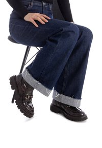 Ciemnoniebieskie jeansy o szerokich nogawkach z postrzępionym jasnoniebieskim mankietem, w połączeniu z błyszczącymi czarnymi butami na obcasie z teksturowanym wykończeniem i detalem zapięcia.