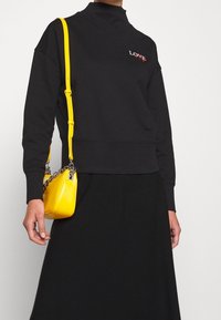 Sweat-shirt noir à col montant avec une broderie rouge "LOVE" ; associé à un petit sac à main en cuir jaune avec une bandoulière chaîne argentée.