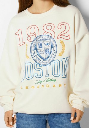 Cream sweatshirt with red "1982," blue "Boston," yellow "Legendary," and a blue athletic crest with "Make Your Own Luck" text.
