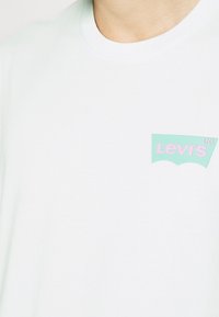 T-shirt blanc à col rond avec un petit logo Levi's vert menthe et rose sur la poitrine gauche, porté par une personne.