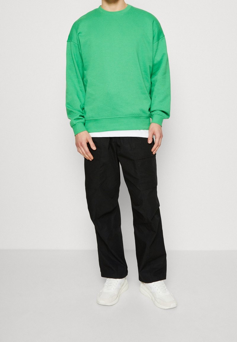 Sudadera verde de algodón con mangas largas, cuello redondo y puños acanalados, combinada con pantalones cargo negros y zapatillas blancas.